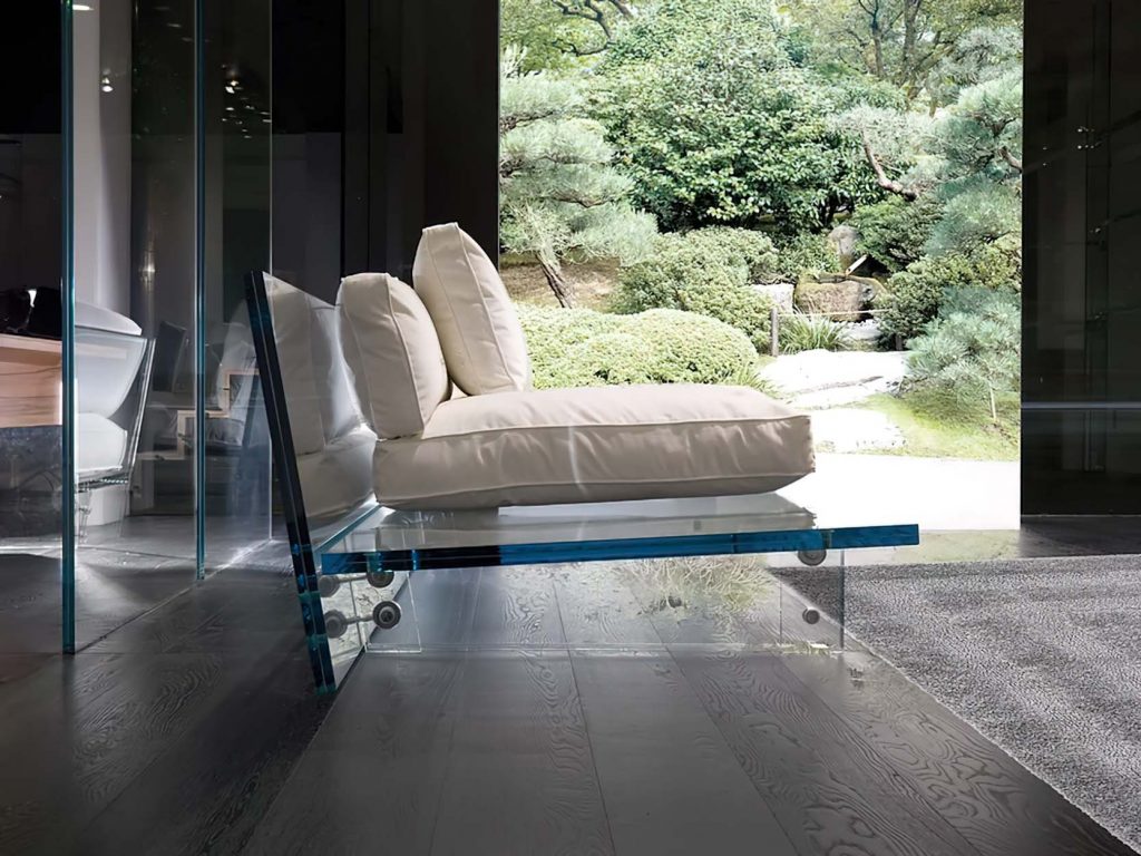 A contemporary beige sofa with plush cushions on a transparent glass base, positioned indoors with a serene garden view through floor-to-ceiling glass walls