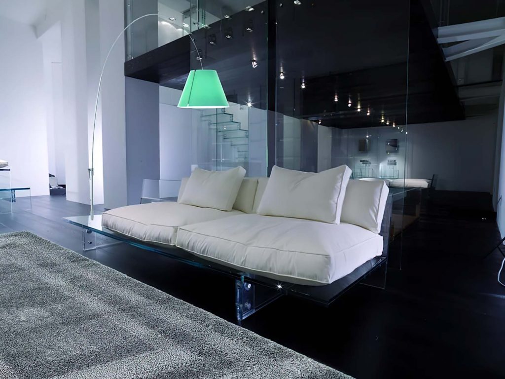 A modern white sofa with plush cushions on a transparent glass base, set in a sleek, dark interior with reflective surfaces and ambient lighting