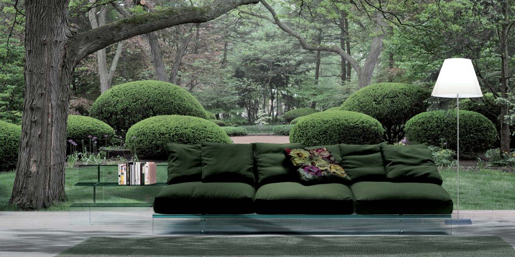 A luxurious green sofa with plush cushions on a transparent glass base, seamlessly blending into a lush garden setting with manicured greenery in the background