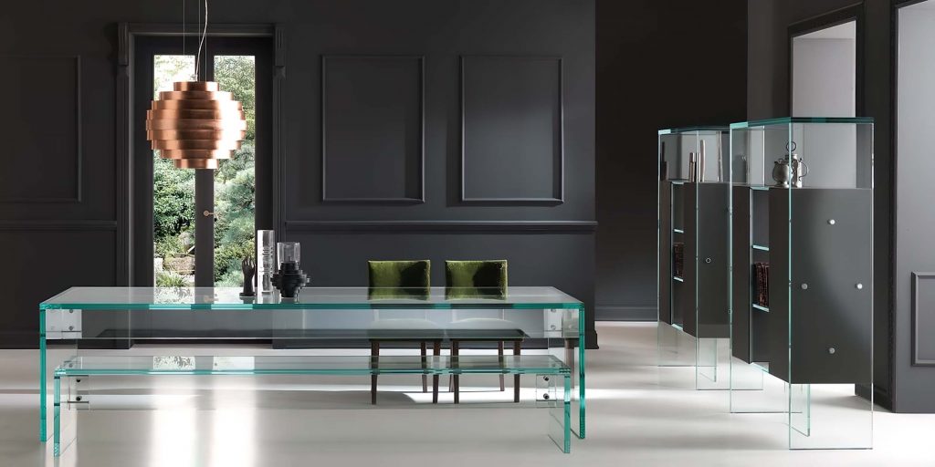 A contemporary glass dining table with a sleek transparent design, set in a modern interior with dark walls and green velvet cushions on the chairs
