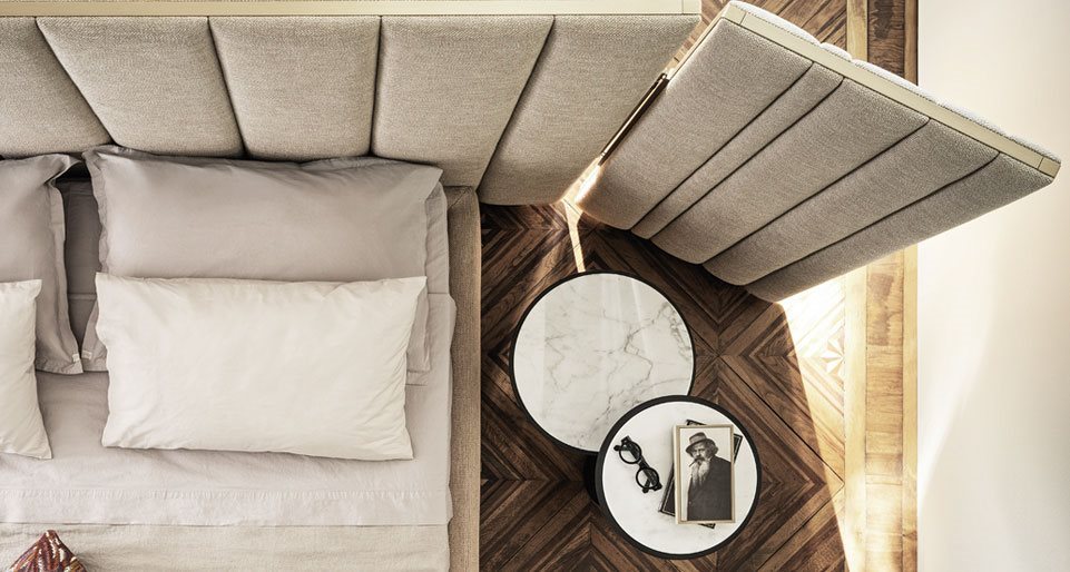 Yuki Fante bedside table with a round marble top next to a plush upholstered bed, set on a herringbone wood floor for a sophisticated and modern bedroom aesthetic