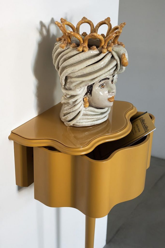 Carlino side table in gold color. Top with drawer and central leg against a white wall.