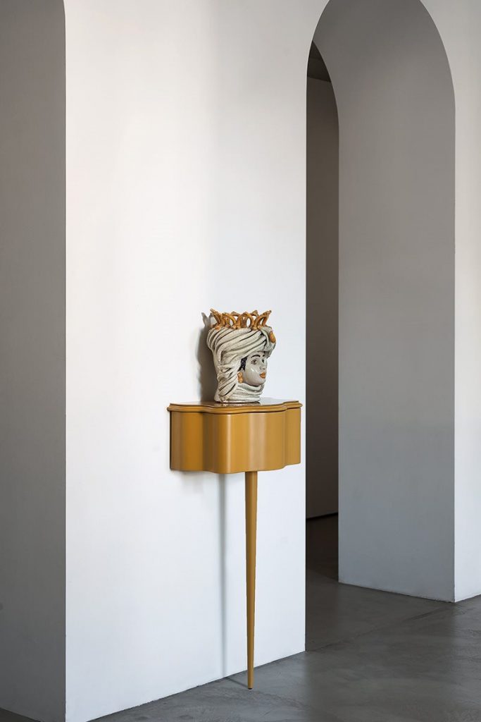 Carlino side table in gold color. Top with drawer and central leg against a white wall.