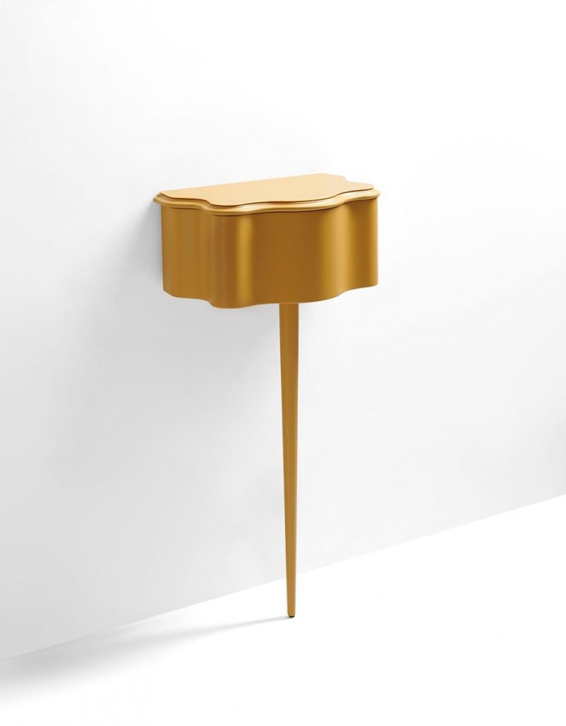 Carlino side table in gold color. Top with drawer and central leg against a white wall.