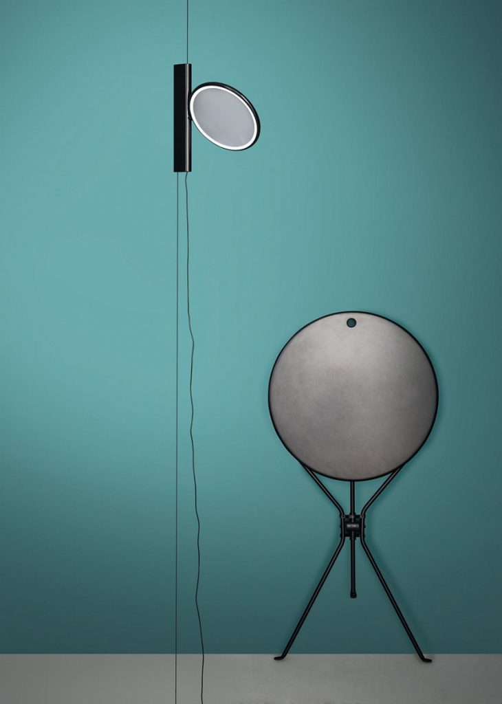 Cumano black side table with a minimalist design, featuring a round top and a slender metal base on a gray floor and next to a blue wall.