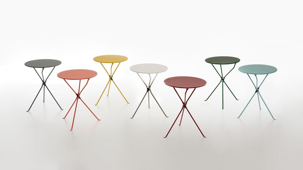Seven Cumano side tables with a round top and a slender metal base, one in white, one in black, one in orange, one in yellow, one in green, one in blue and one in red on a white background.