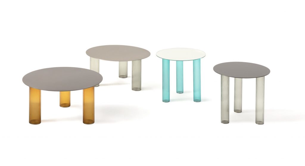 Four Echino coffee tables. Round gray top and three semi transparent cilindrical legs, one in bronze, two in gray and one in blue on a white background.