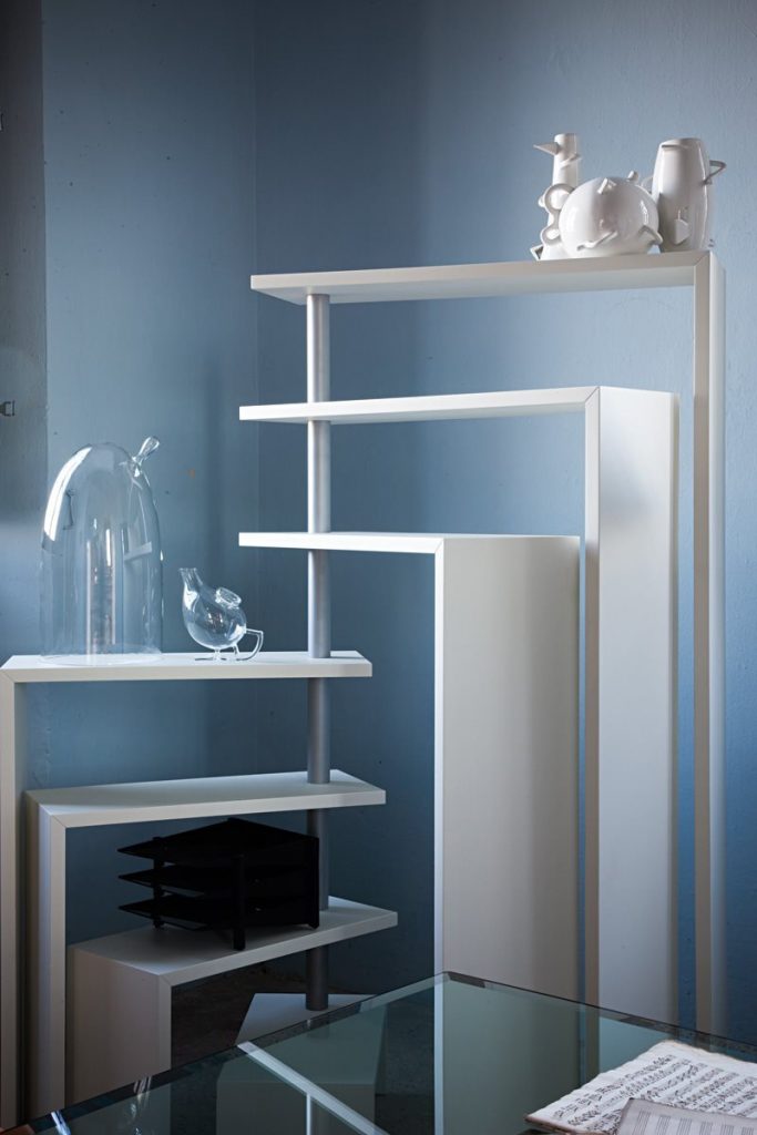 White Joy bookcase with seven moveable shelves around a central tube in front of a blue wall.
