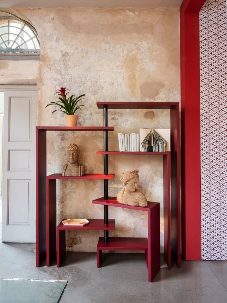 Red Joy bookcase with seven moveable shelves around a central tube on a gray floor and yellow wall.