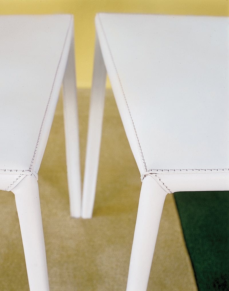 Closeup of two Lea chairs featuring a high backrest and a upholstered seat and backrest in white fabric with four white legs on a rug.