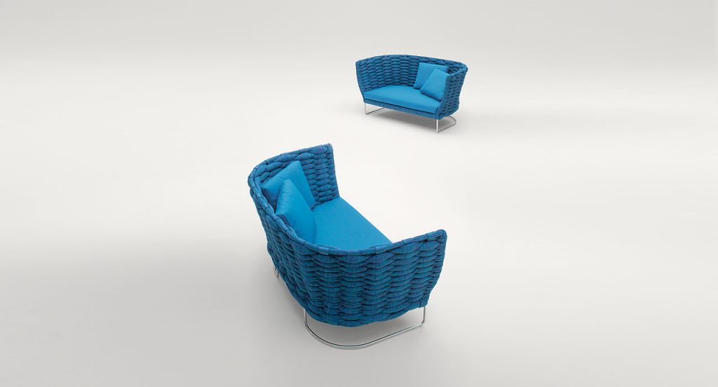 Closeup of Ami Indoor Sofa: Two blue upholstered sofas with woven sides and curved backrest with two silver legs on a white background.