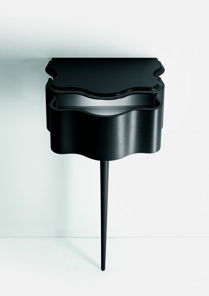 Carlino side table in black color. Top with drawer and central leg against a white wall.