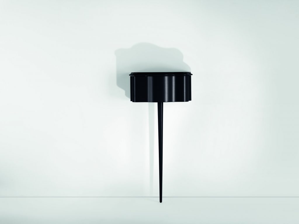 Carlino side table in black color. Top with drawer and central leg against a white wall.