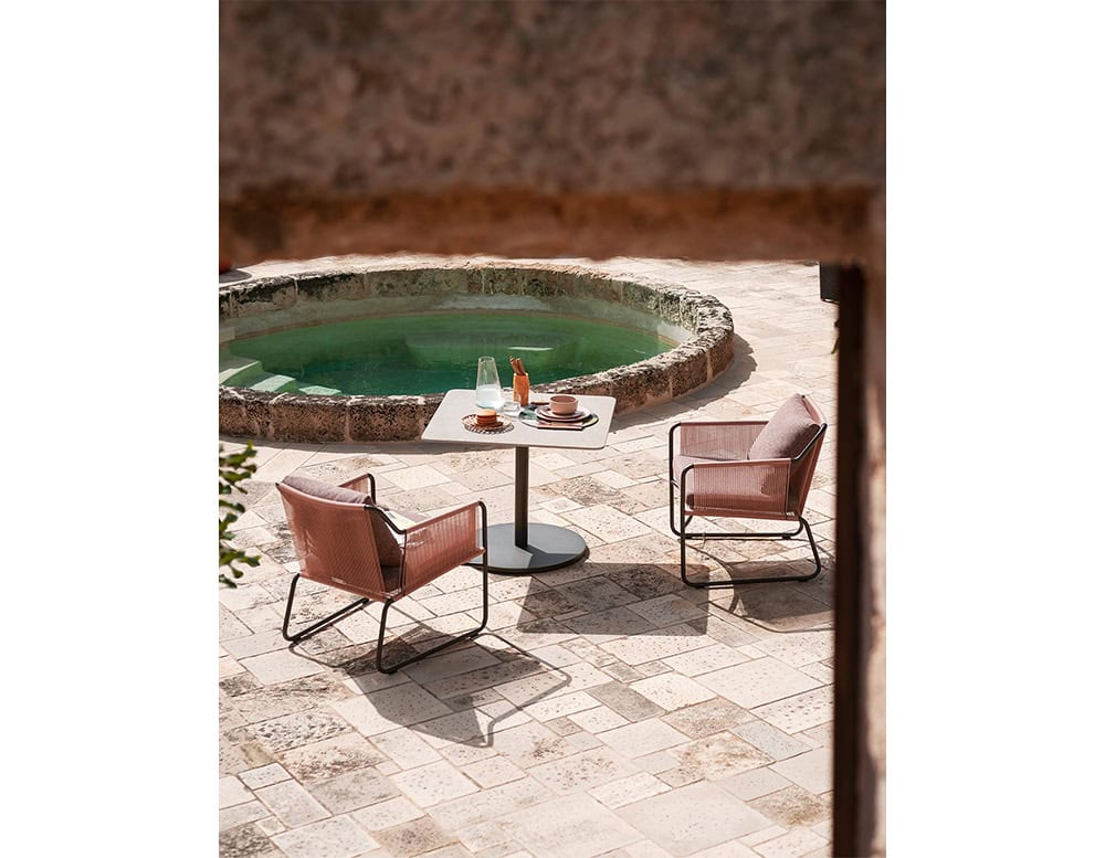 A square outdoor coffee table with a light grey top and black base is set between two pink woven chairs on a stone terrace beside a round plunge pool.