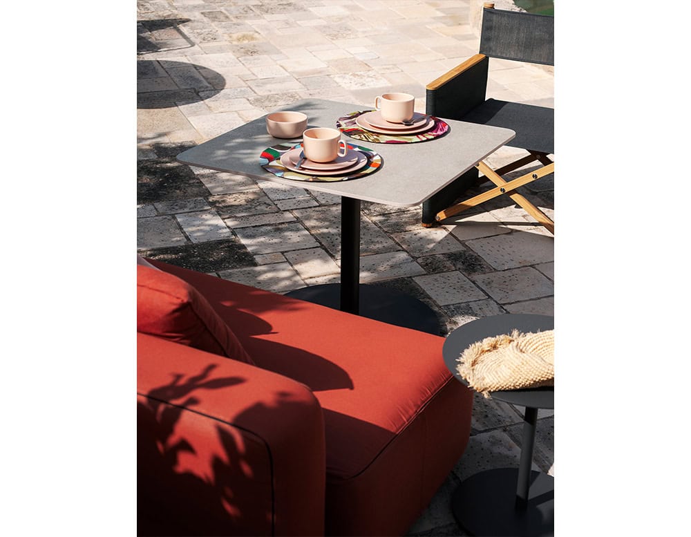 A square outdoor coffee table with a light grey top and black pedestal base, styled with colorful tableware and surrounded by lounge seating on a sunlit patio.