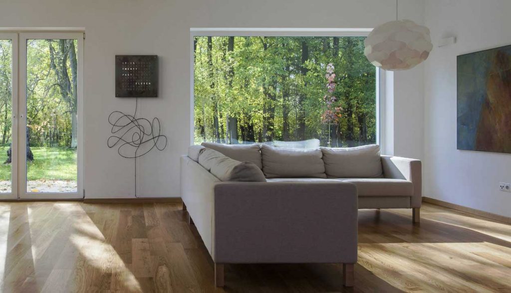 QLOCKTWO Classic in black on a white wall with a sofa in front with a tv to the right wall and windows to the left and right of the clock