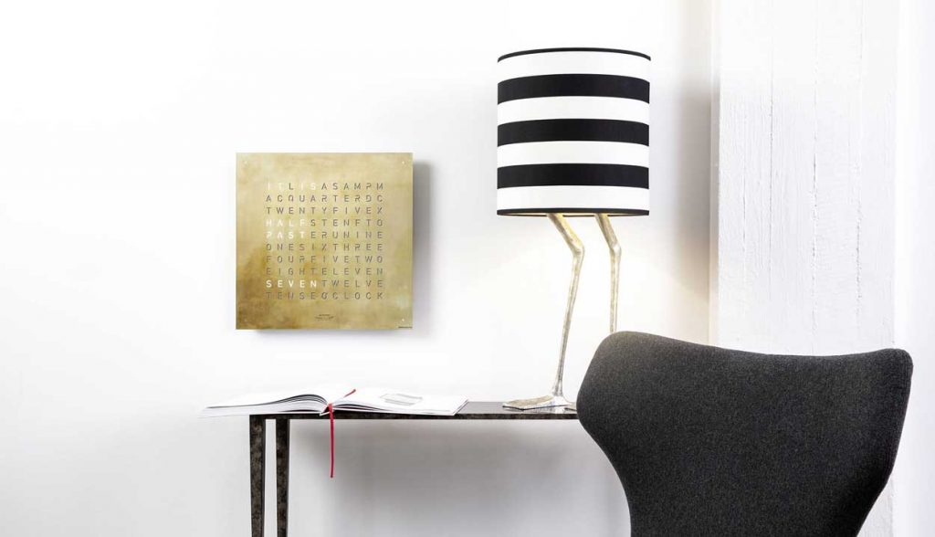 QLOCKTWO Classic in gold on a white wall with a desk in front and a lamp on the desk