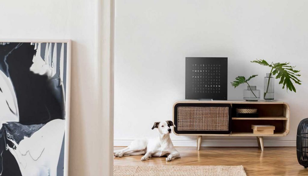 QLOCKTWO Classic in black hanging on a white wall on top of a desk with a dog next to the desk