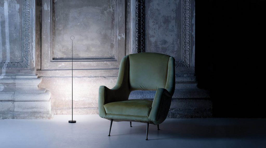Anima light shining against a renaissance looking wall with an army green chair to the right of the light