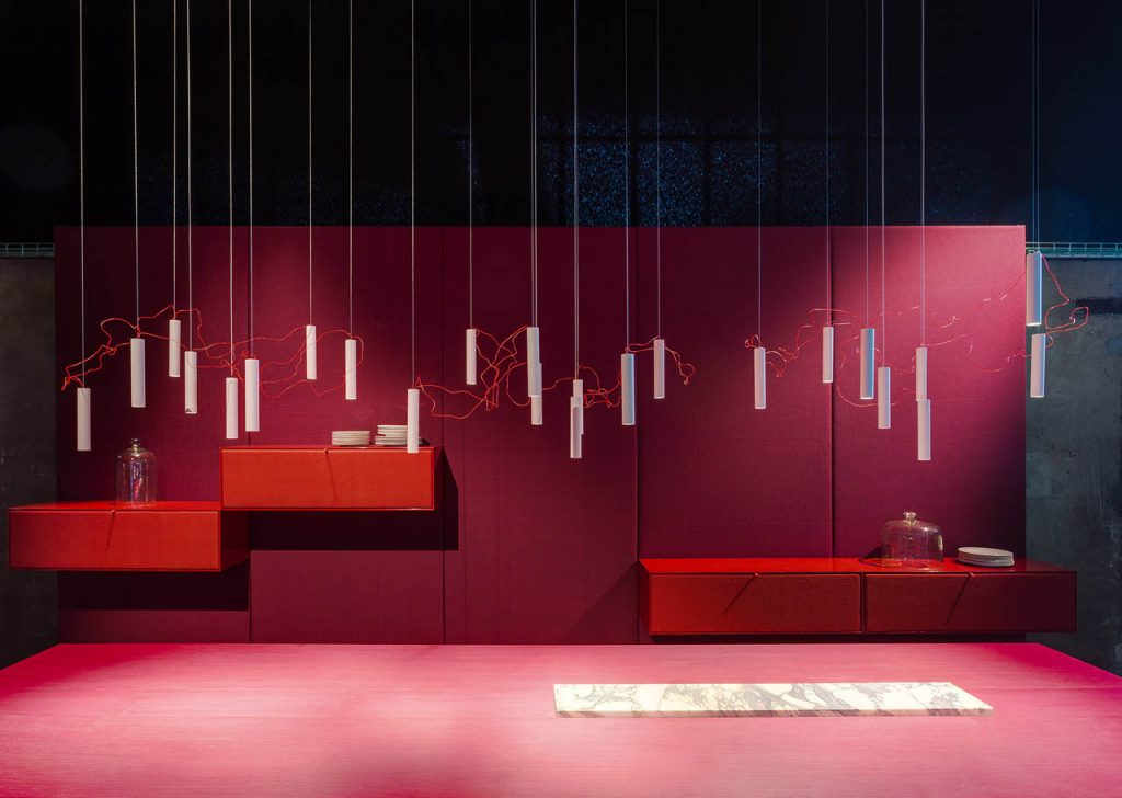 Simbiosi chandelier hanging in front of a red wall