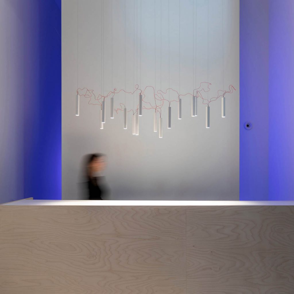 Simbiosi chandelier hanging in front of a white wall with a person walking underneath