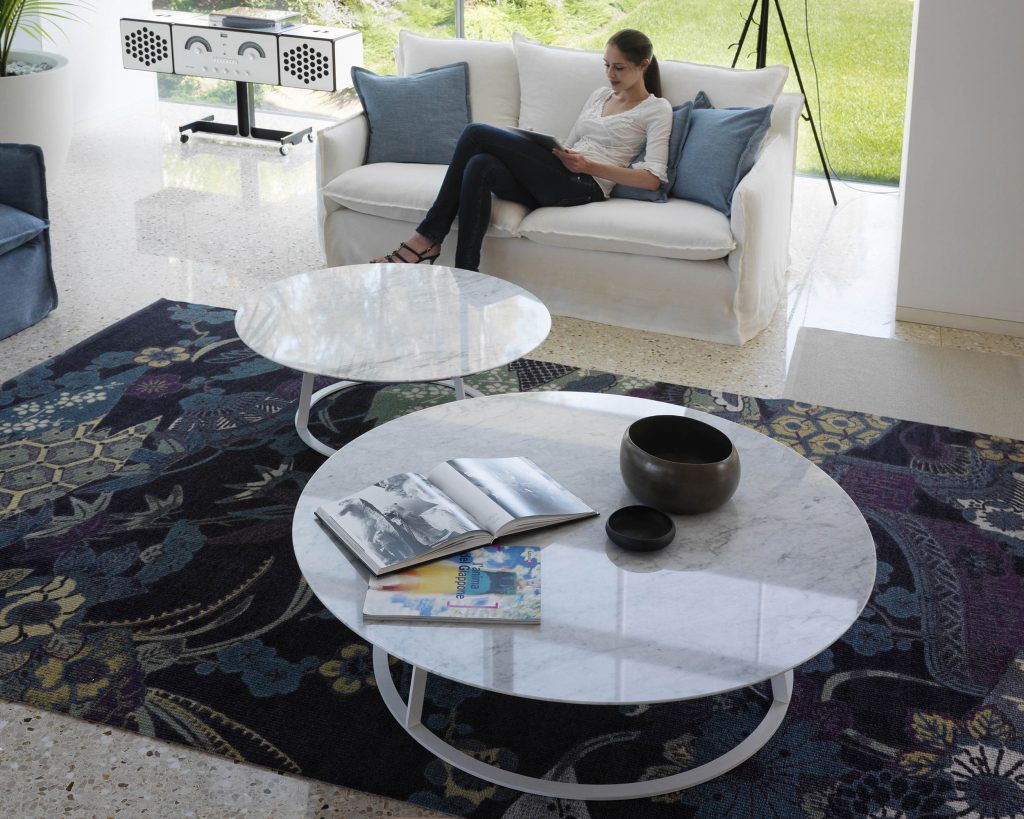 Albino Family wide white top in front of a smaller version in front of a cream colored sofa on top of a blue carpet with a person sitting on the sofa