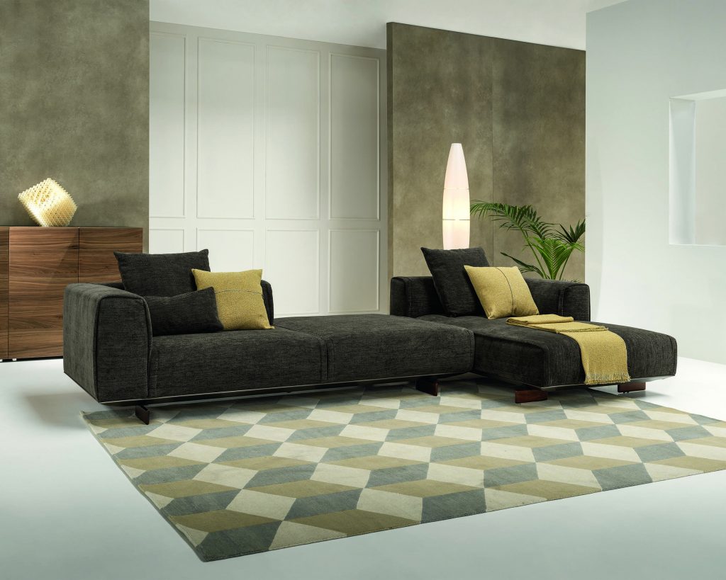Dark grey Coleman with the right side having a yellow thick stripe going through the middle. The Coleman is on top of a optical illusion pattern rug with white walls surrounding the sofa
