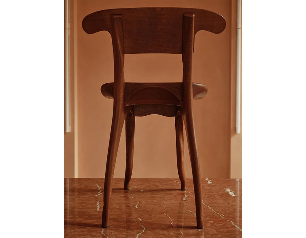 The BD Batllo Chair displays elegant handcrafted detailing, with sculpted legs and a curved backrest. The piece is crafted in a deep, warm brown wood, emphasizing its organic silhouette and artisanal character.