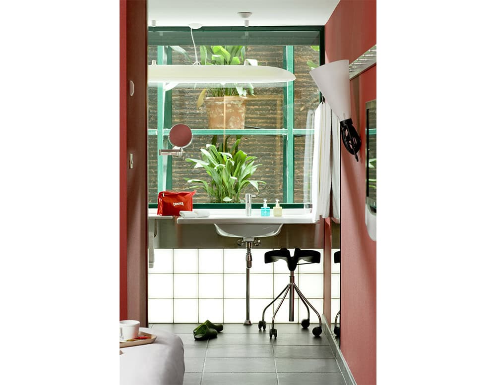 The BD Binaria Stool is placed in a modern bathroom space. Its black saddle-style seat and polished chrome base with wheels add a sleek, functional touch to the bright interior with red walls, glass elements, and natural greenery.