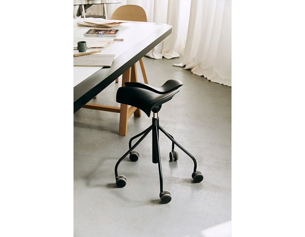 The BD Binaria Stool features a sleek black saddle-style seat with an ergonomic curve, supported by a five-spoke base with smooth caster wheels. Its contemporary design pairs well with creative and collaborative workspaces.