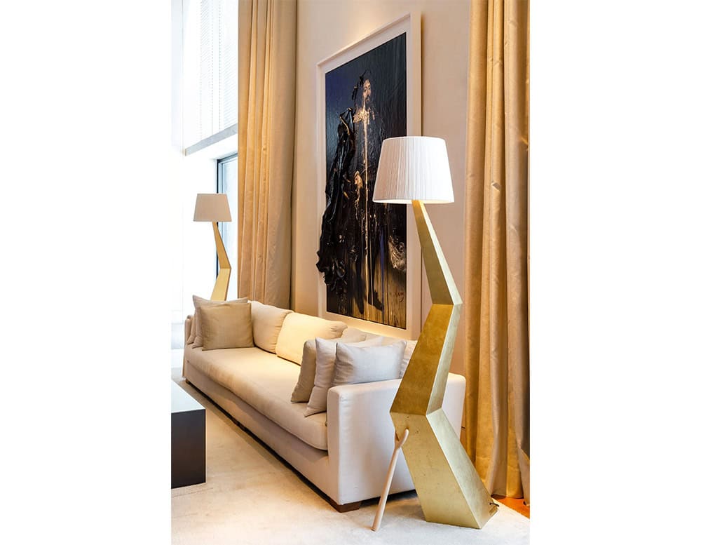The lamp’s gold angular base and white shade bring sculptural elegance to this cozy living room. Positioned beside a neutral sofa, it complements the soft beige curtains and adds a bold, geometric contrast to the space.