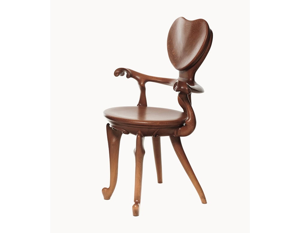 The BD Calvet Armchair is shown at a three-quarter angle, revealing the sculptural backrest, curved armrests, and rounded seat. Crafted in warm brown wood, the piece showcases smooth surfaces and visible natural grain.