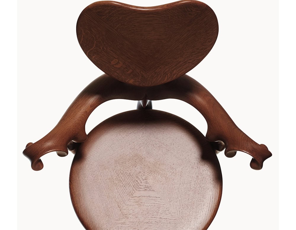 The BD Calvet Armchair is shown from above, highlighting its heart-shaped backrest, curved arms, and round seat. The polished oak wood displays deep reddish-brown tones with visible natural grain patterns.