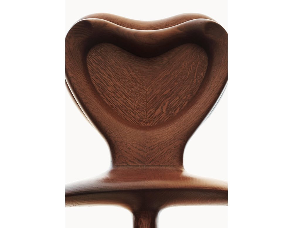 The BD Calvet Armchair features a heart-shaped backrest in richly grained oak. This close-up highlights the smooth carving and the flowing, organic lines. The warm brown tones of the wood emphasize its natural texture.