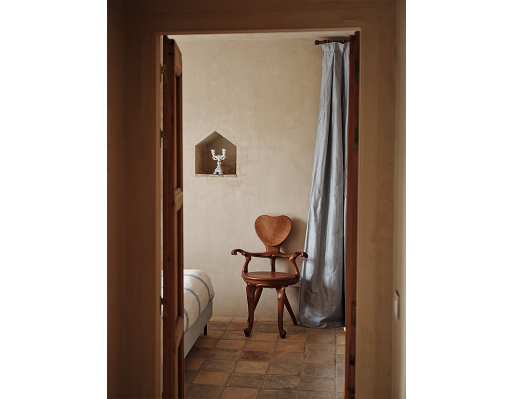 The BD Calvet Armchair sits gracefully in a serene bedroom setting. Its sculptural wooden structure with curved armrests and heart-shaped backrest stands out as a refined design element within the calm, rustic ambiance.