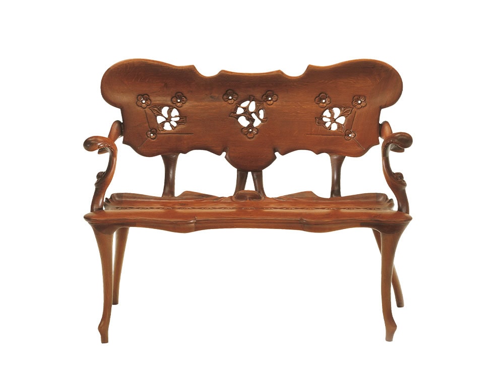 The BD Calvet Bench features an ornate carved backrest adorned with floral motifs. Its sinuous legs and flowing lines highlight a craftsmanship that merges structure with decorative artistry.