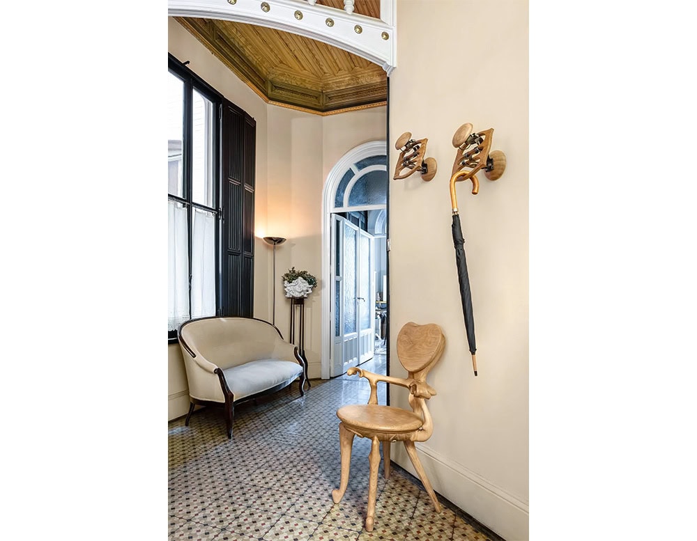 An inviting entrance area highlights the BD Calvet Hanger, mounted elegantly on the wall. The space also features a wood chair with an organic silhouette, a classic settee, and mosaic-tiled flooring that adds a historic charm.