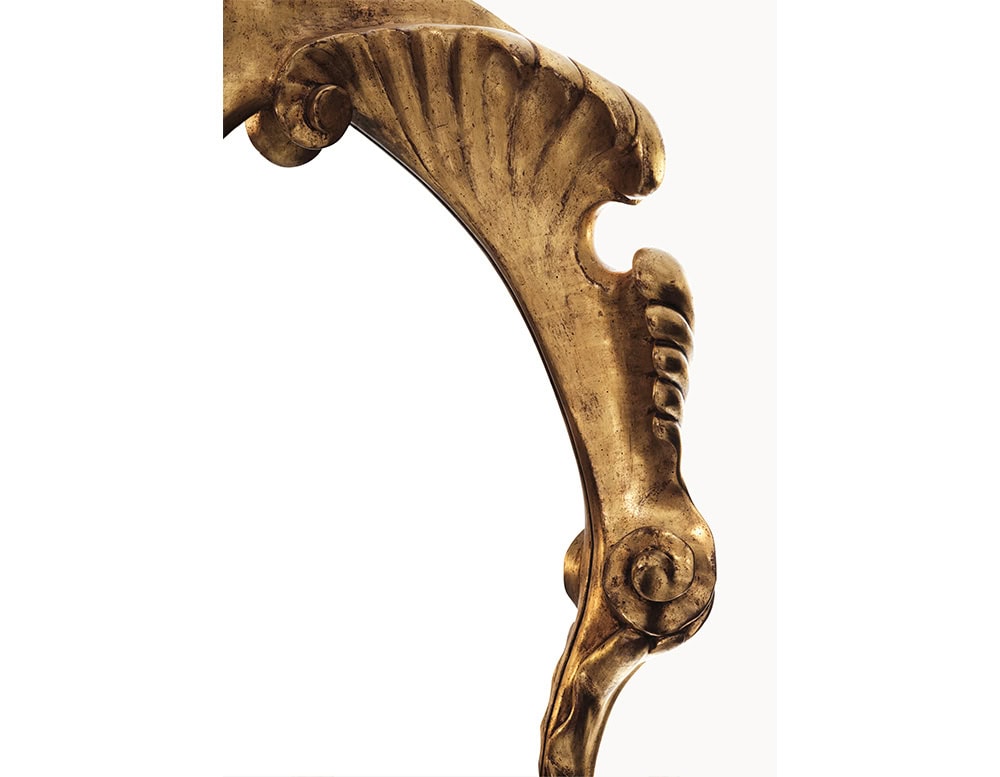 A close-up of the BD Calvet Mirror reveals its elaborate hand-carved wooden frame, finished in radiant gold leaf. The design highlights flowing curves and intricate textures that reflect the mirror's distinctive artisanal quality.