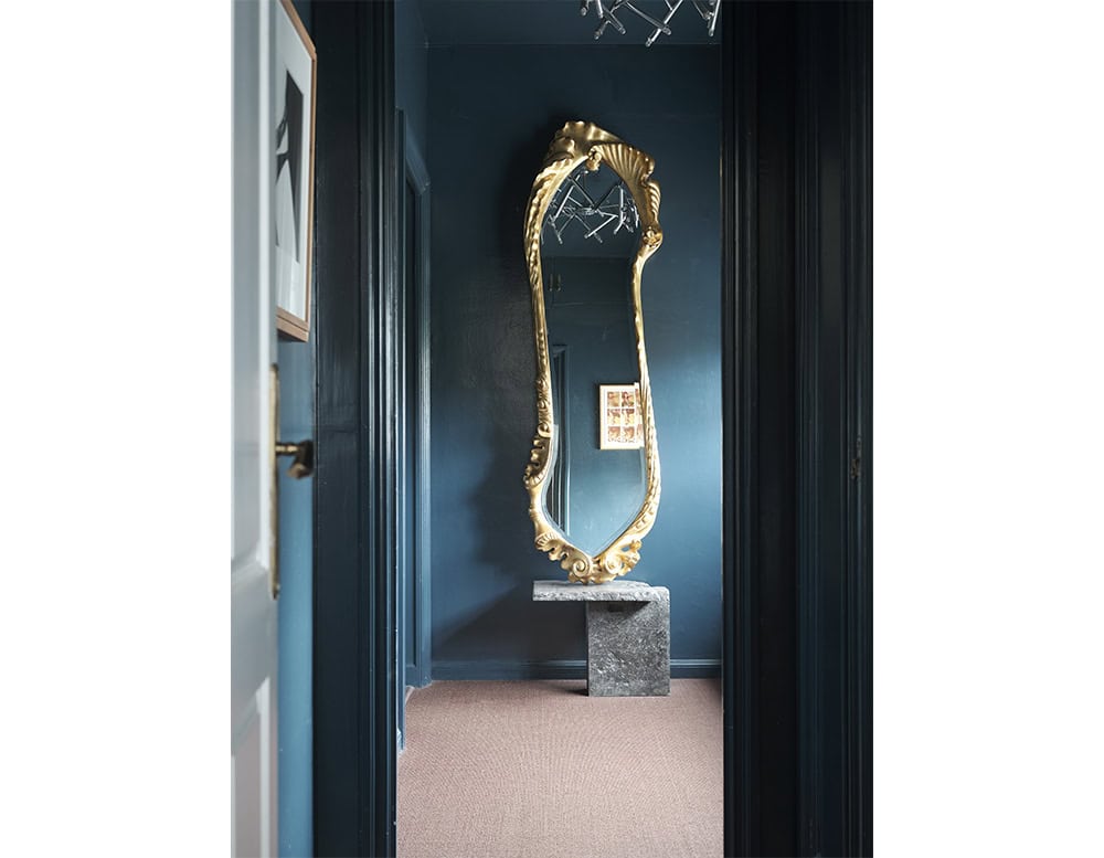 The BD Calvet Mirror features a lavish hand-carved wooden frame with fluid, sculptural lines. Its gold leaf finish adds opulence, reflecting softly in a moody hallway with dark walls, a stone pedestal, and warm flooring.