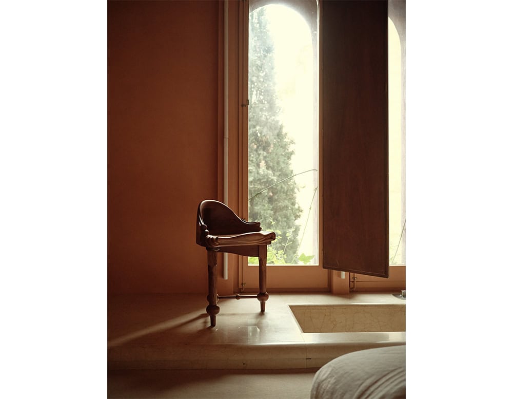 A sculptural wooden stool sits near a tall window, bathed in soft natural light. Its rich wood texture contrasts with the smooth beige floors and walls, creating an intimate, serene corner with a sculptural presence.