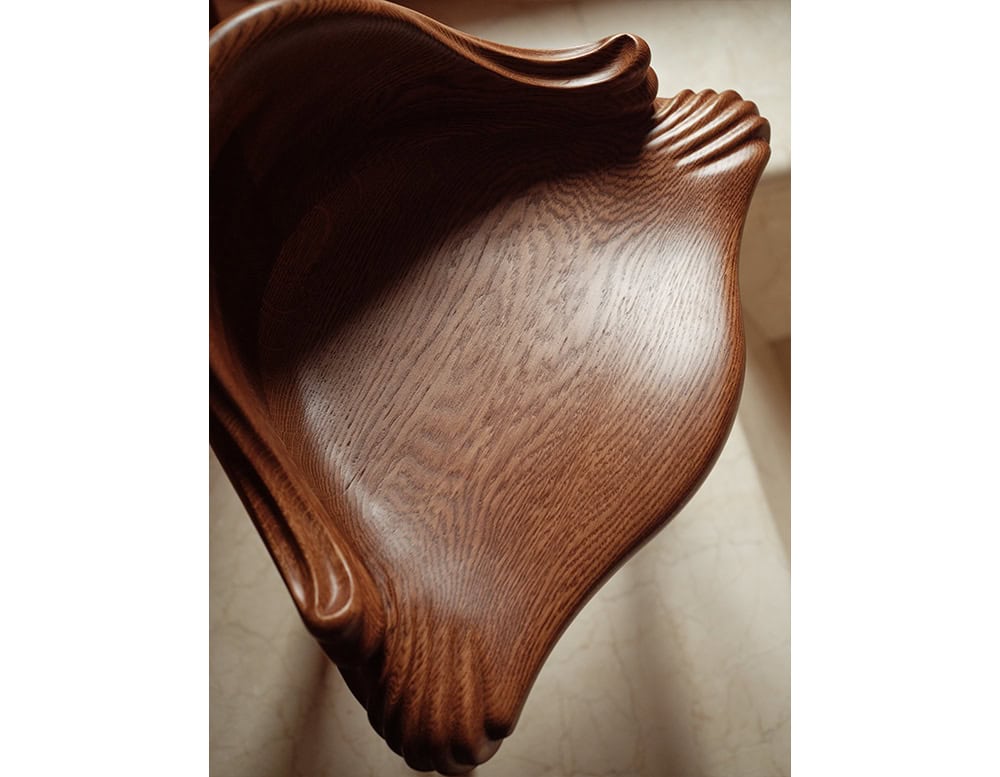 A close-up of the BD Calvet Stool highlights its rich, sculpted oak seat with a smooth, polished surface. The flowing carved details resemble organic folds, accentuating the natural grain of the wood with a warm brown finish.