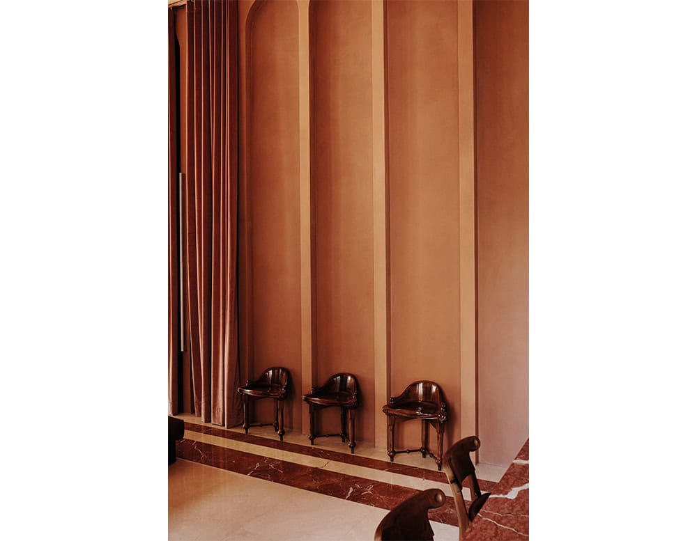 Three Calvet Stools sit against a paneled wall in a warm terracotta tone. The stools have a sculptural wooden form with curved lines and rich dark polish, contrasting the smooth vertical geometry of the architectural backdrop.