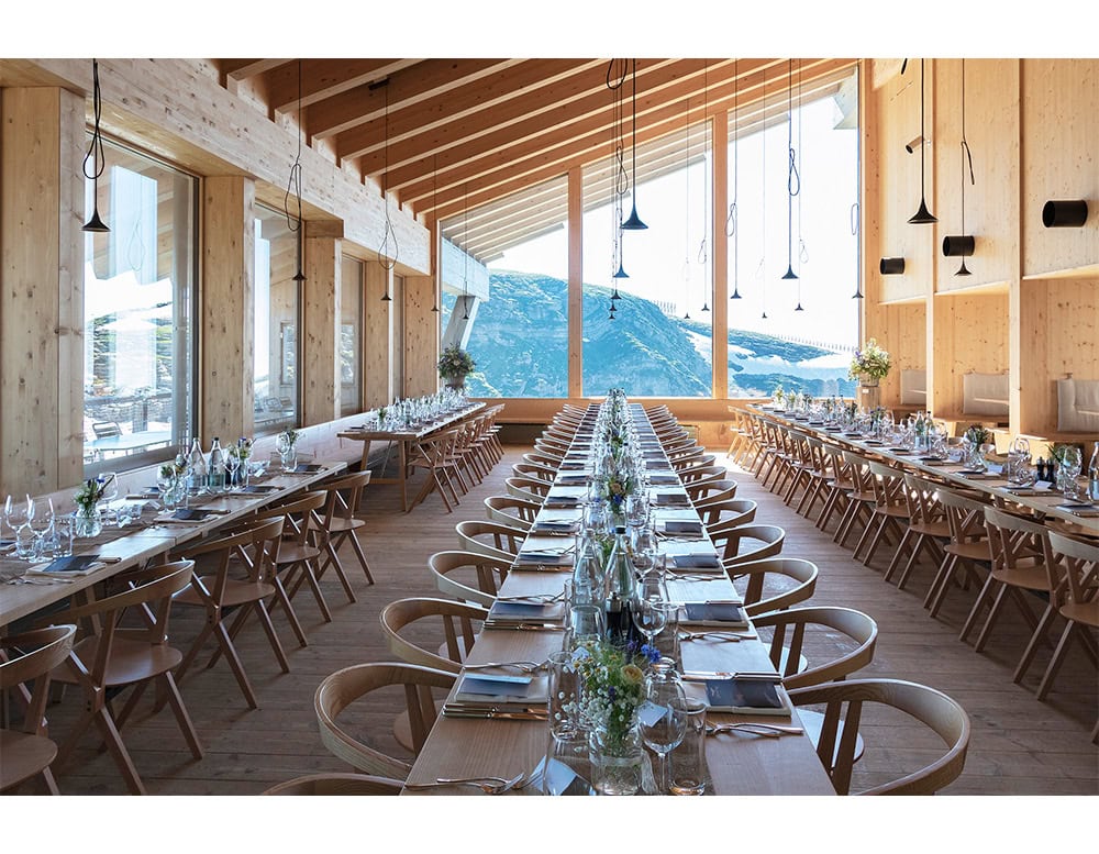 An alpine dining room with BD Chair B in natural wood. The chairs’ curved backs echo the exposed beams, while large windows frame breathtaking mountain views, blending architecture and nature.