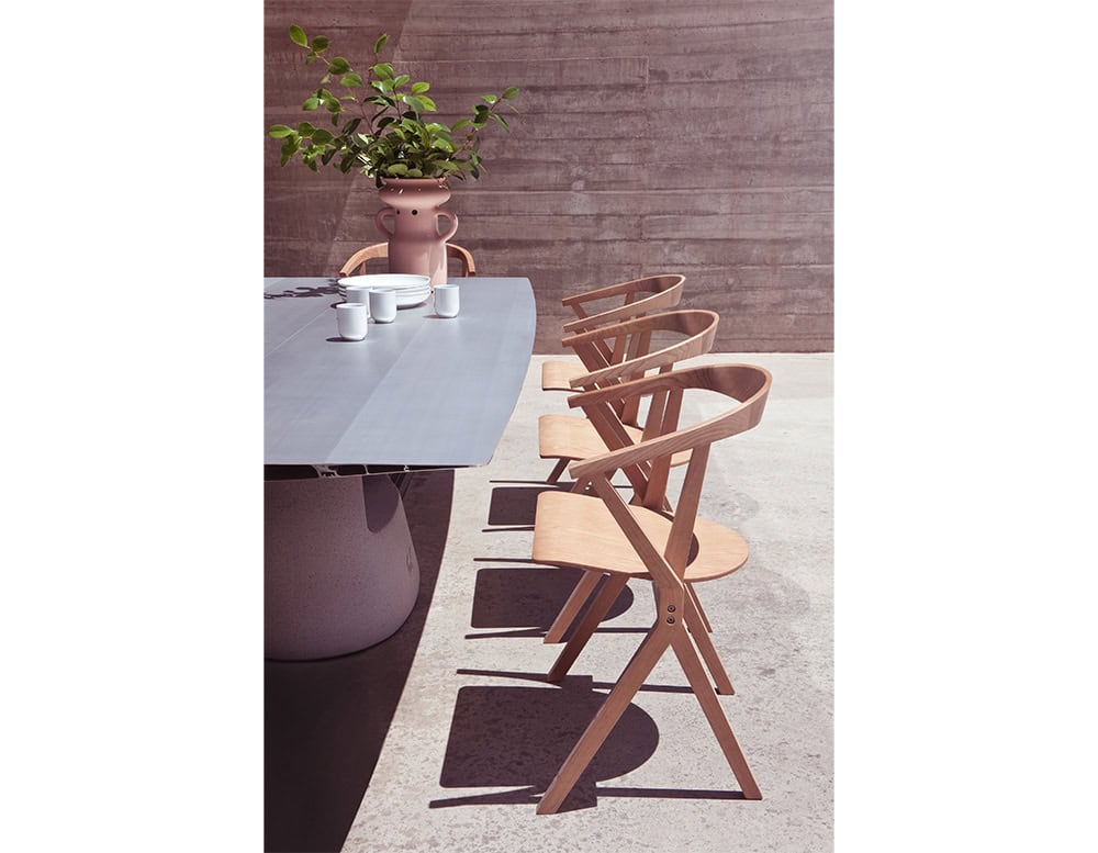 A sunlit outdoor dining scene featuring the BD Chair B in natural wood finish. The sculptural simplicity of the chairs complements the sleek grey table, while a playful ceramic vase with greenery adds a whimsical touch to the composition.