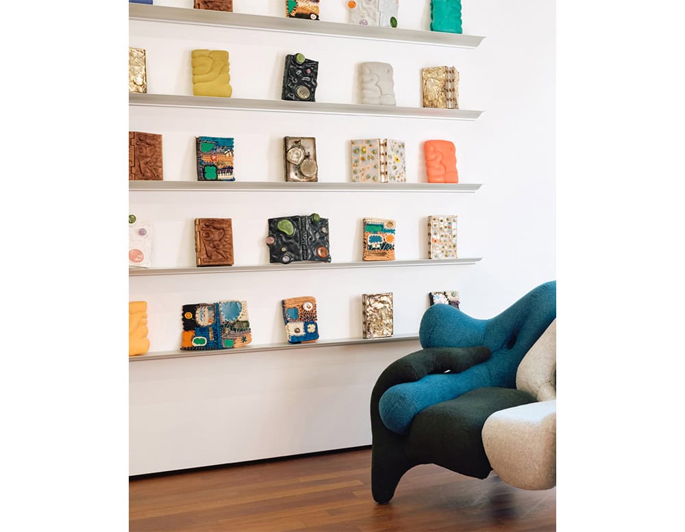 The BD Cornisa Shelf is shown here mounted on a white wall, displaying a colorful collection of sculptural art books and objects, creating a vibrant, gallery-like arrangement next to an upholstered lounge chair.