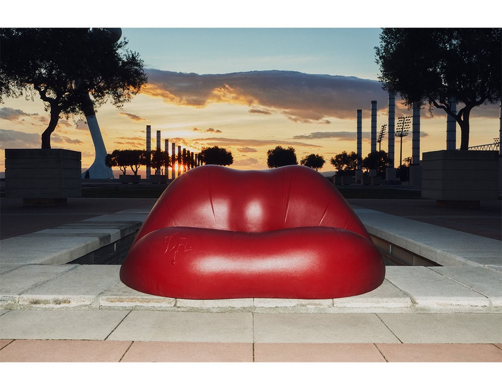 The Dalilips Sofa sits boldly in an urban setting at sunset. Its striking red lip-shaped form creates a surreal contrast against the geometric lines of the plaza and the warm sky in the background.