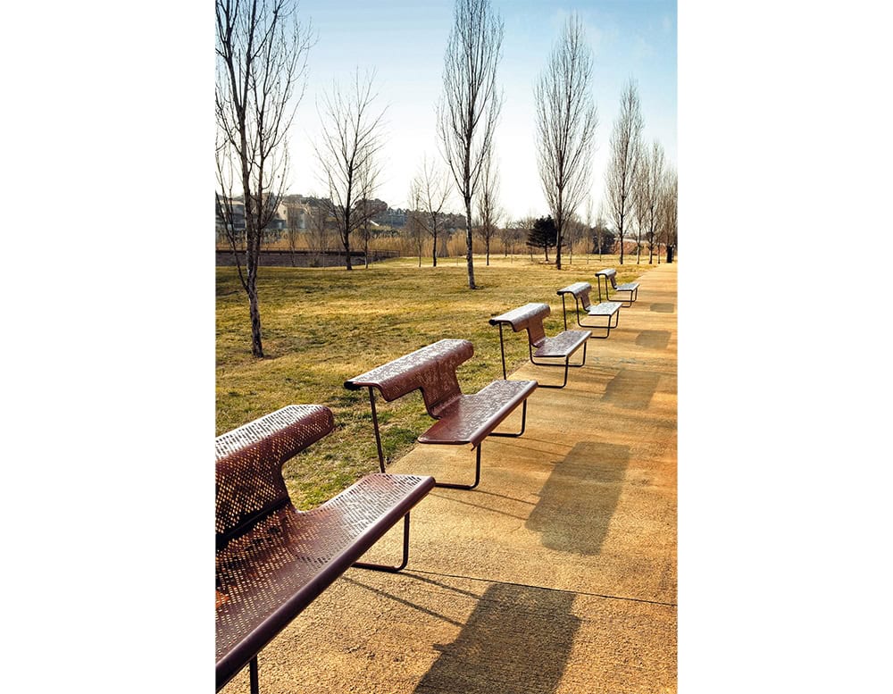 A row of El Poeta benches lines the park walkway, featuring perforated metal surfaces and a sculptural design. Each bench integrates seat and backrest elements, offering comfortable outdoor seating while maintaining visual openness.