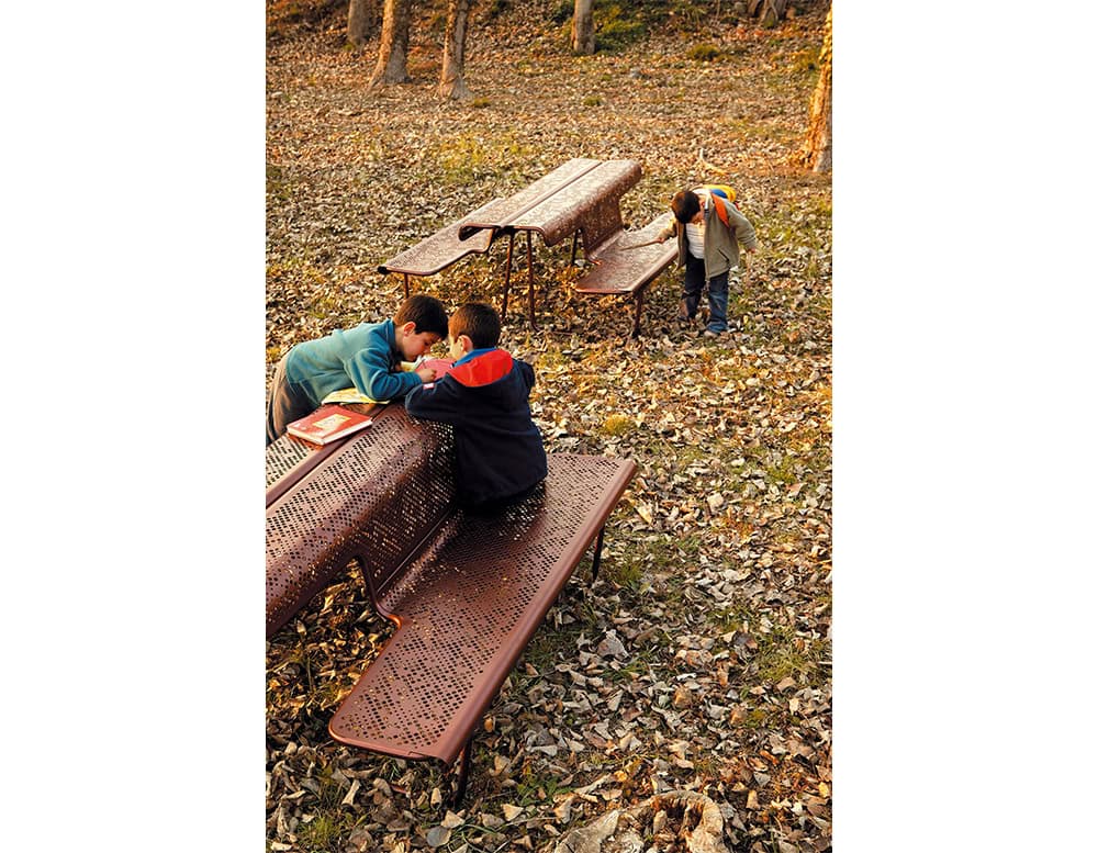 The El Poeta benches are shown in a park setting, featuring perforated metal surfaces with curved edges and integrated seats. Their continuous design creates both bench and tabletop surfaces, encouraging gathering and interaction in outdoor spaces.