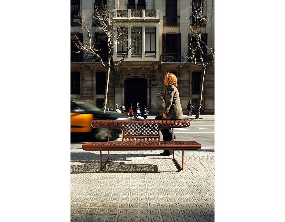 The El Poeta bench features a perforated metal backrest and seat with rounded edges, offering an open design that blends into the urban landscape. Its slender metal legs provide a stable yet minimal support structure, complementing city sidewalks.