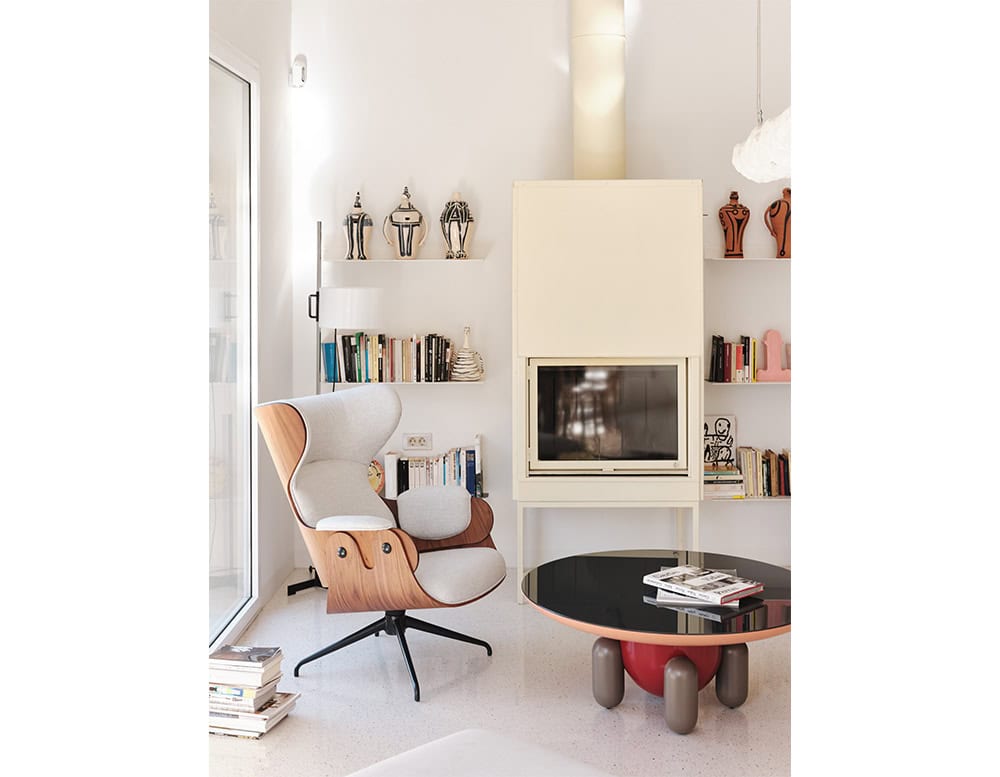 A round coffee table with a glossy black top and coral edge, supported by playful rounded legs. Nearby, a sculptural wood lounge chair sits by a wall of bookshelves, with modern ceramic vases and graphic art adding visual interest.